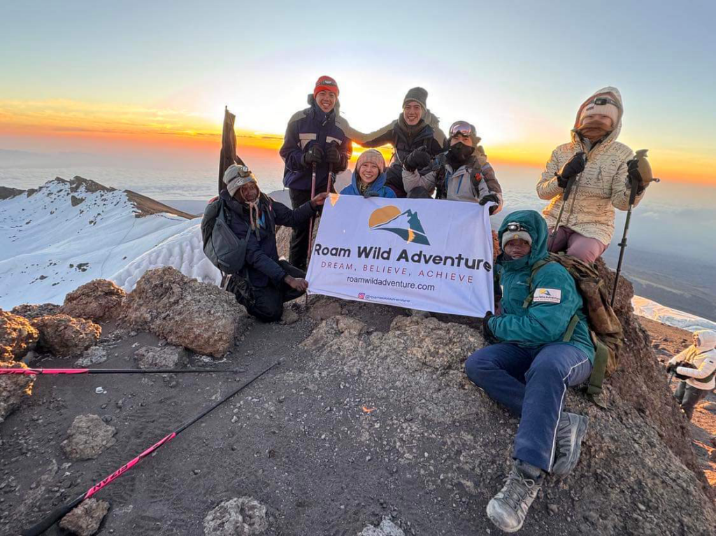 Group of hikers holding a sign for Roam Wild Adventure atop Mt. Kilimanjaro.