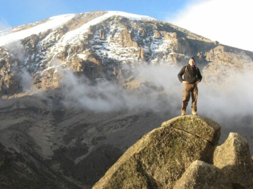 A person standing on a rock in front of the view of the summit of Mount Kilimanjaro.