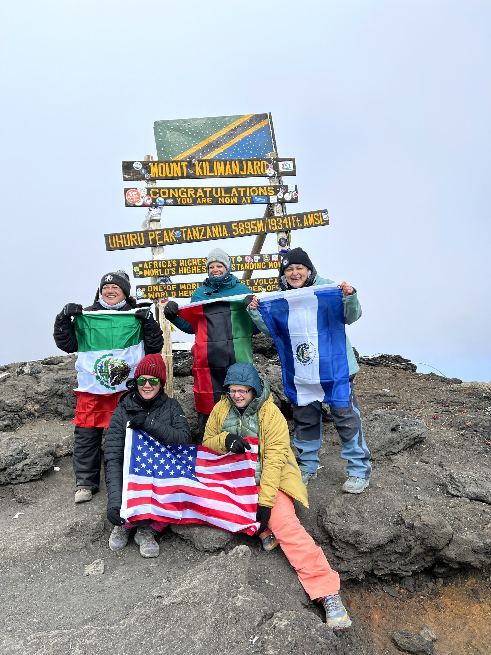 A group of women holding flags and signs at the summit of Mount Kilimanjaro.